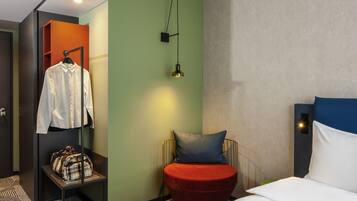 Une chambre d’hôtel avec un lit, une chaise rouge, une petite table sur laquelle se trouve un sac à main, un manteau accroché et une lampe fixée au mur.