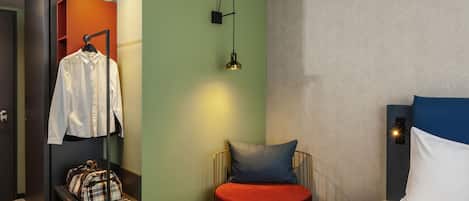 A hotel room with a bed, a red chair, a small table with a handbag, a hanging coat, and a wall-mounted lamp.