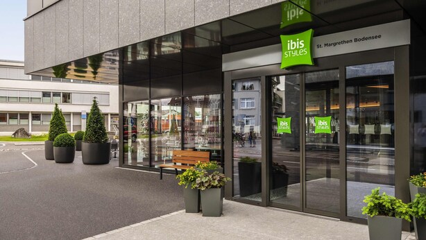 Un edificio moderno con fachada de vidrio, señalización verde con la marca 'ibis Styles' y un banco en el exterior.