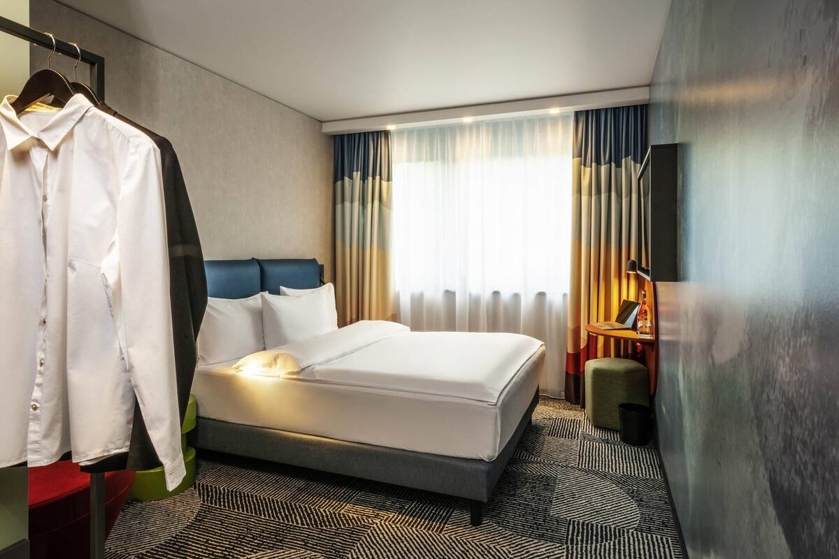 Une chambre d’hôtel comprenant un lit, une armoire, un bureau avec une lampe et une grande fenêtre avec des rideaux.