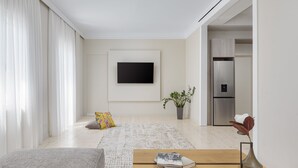 Uma sala de estar moderna com uma televisão de tela plana na parede, uma mesa de centro de madeira com livros, um sofá cinza, um tapete estampado, uma planta em vaso e uma geladeira ao fundo.