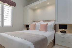 A neatly made bed with white and pink bedding, a bedside table with a lamp, and a window with blinds. - THIRAMENOUS RESIDENCE - ATHENA SUITE with Rooftop Hot Tub (Athina)