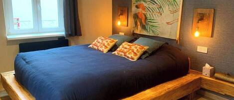 A large bed with a dark blue bedspread and two pillows, a wooden headboard, a painting of a bird on the wall, and two wall-mounted lights.