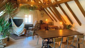 A cozy attic room with wooden beams, a hammock, a dining table, and various chairs. - Charming cottage in the countryside near Strasbourg (Fessenheim-le-Bas)