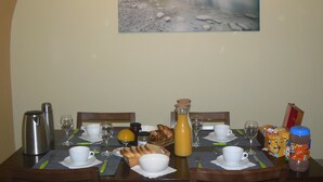 Une table à manger dressée avec des aliments pour le petit-déjeuner, notamment du pain, du beurre et des condiments, dans une pièce dont le mur est orné d’un tableau représentant une montagne.