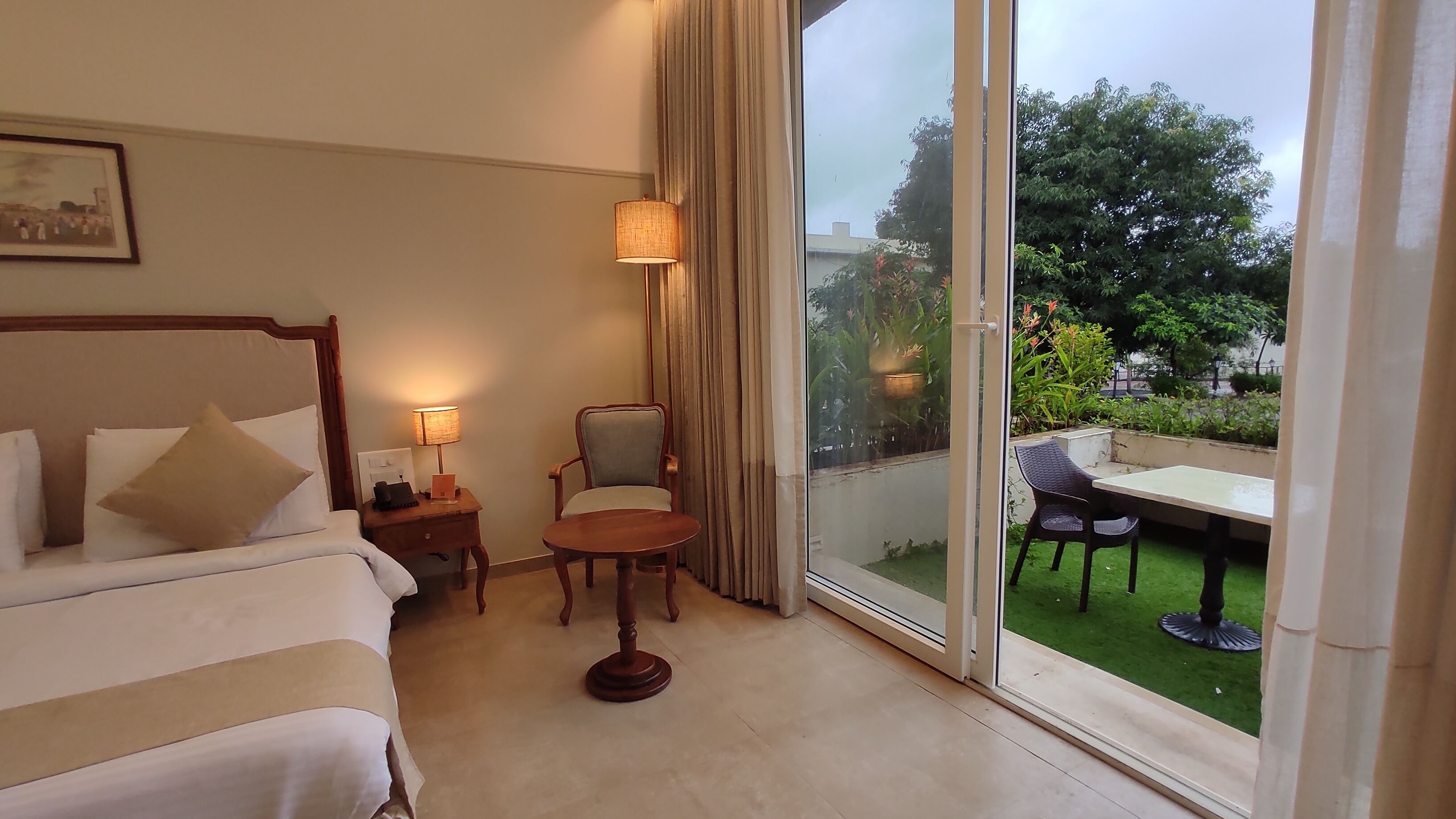 A hotel room with a bed, a chair, a small table, and a view of a garden through a glass door.