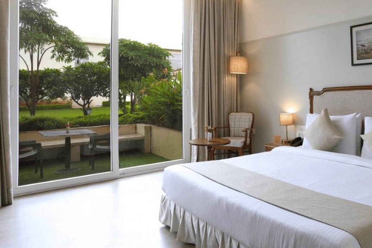 A hotel room with a large bed, two bedside tables, a chair, and a view of a garden through a glass door.