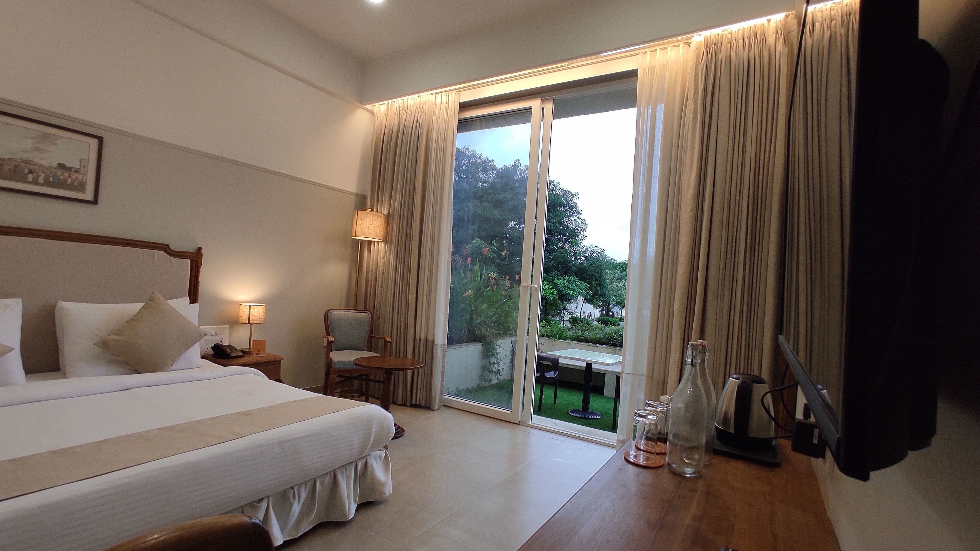 A hotel room with a bed, a chair, a table with a bottle and glasses, and a view through the glass door.