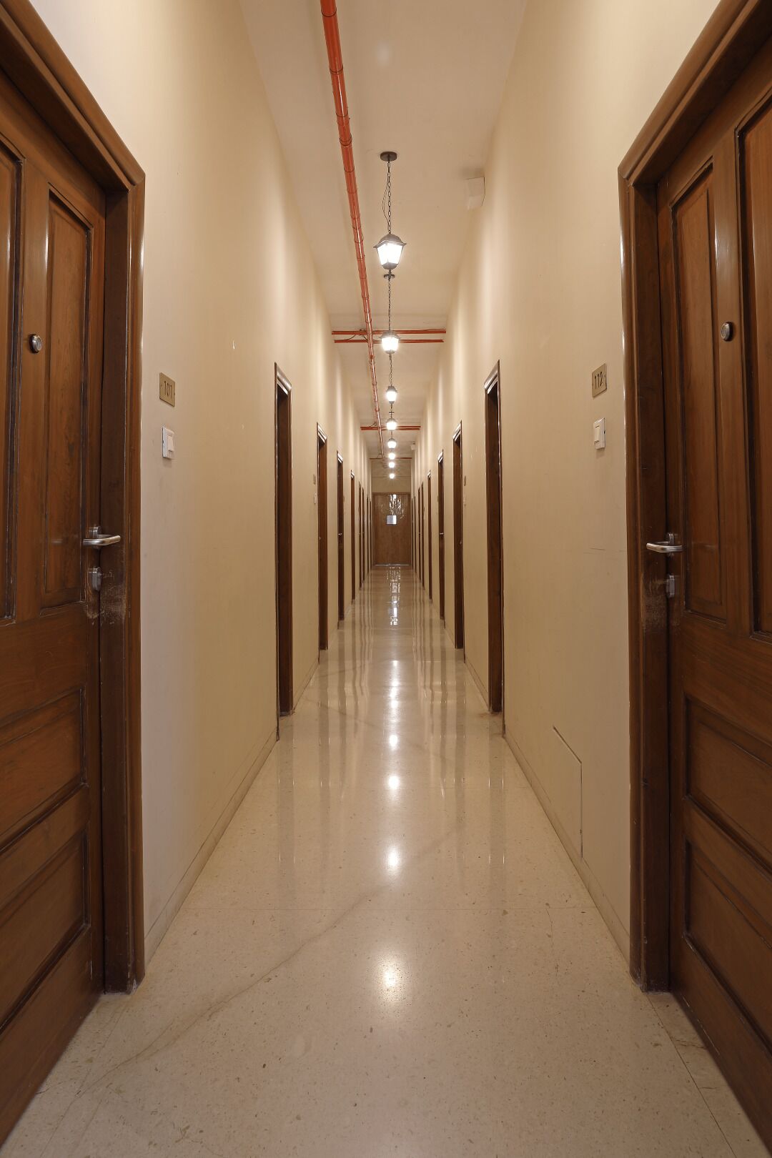 Un largo y estrecho pasillo de hotel con puertas de madera a ambos lados, suelo de baldosas y una serie de luces a lo largo del techo.