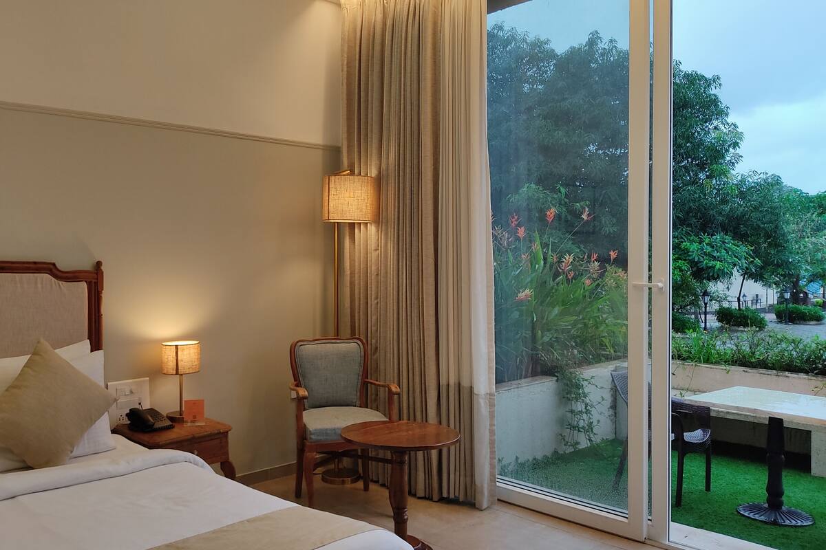 A hotel room with a bed, a chair, a small table, a lamp, and a view of a garden through a large window.