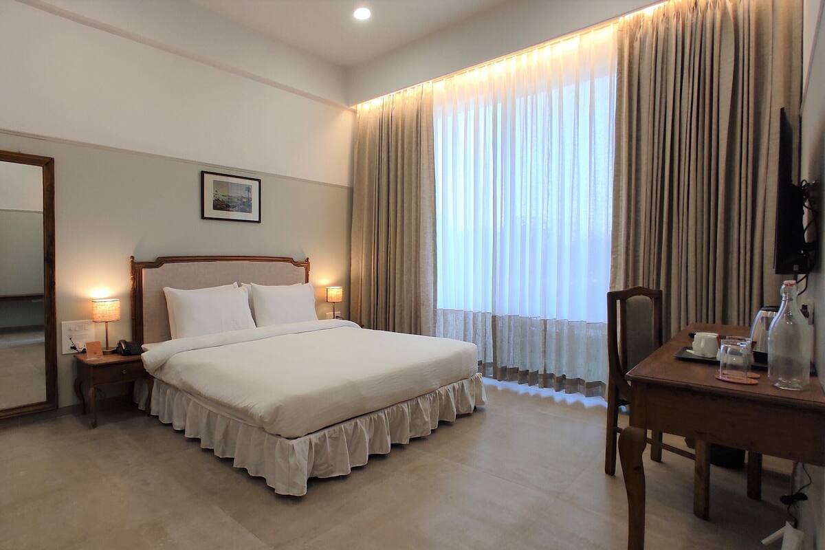 A hotel room with a large bed, a desk with a chair, a mirror, and a window with curtains.