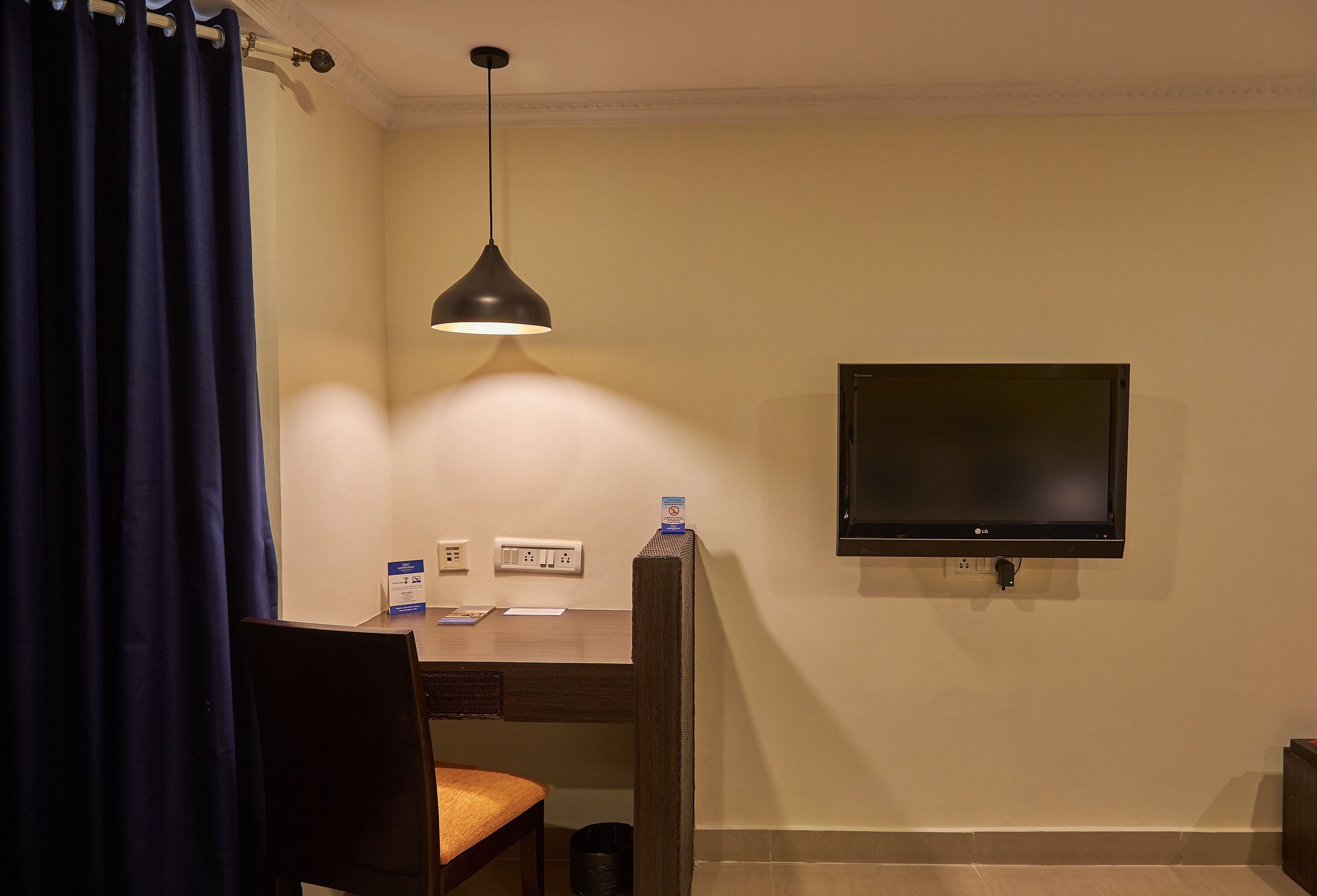 A hotel room with a desk, chair, TV, and a hanging pendant light.
