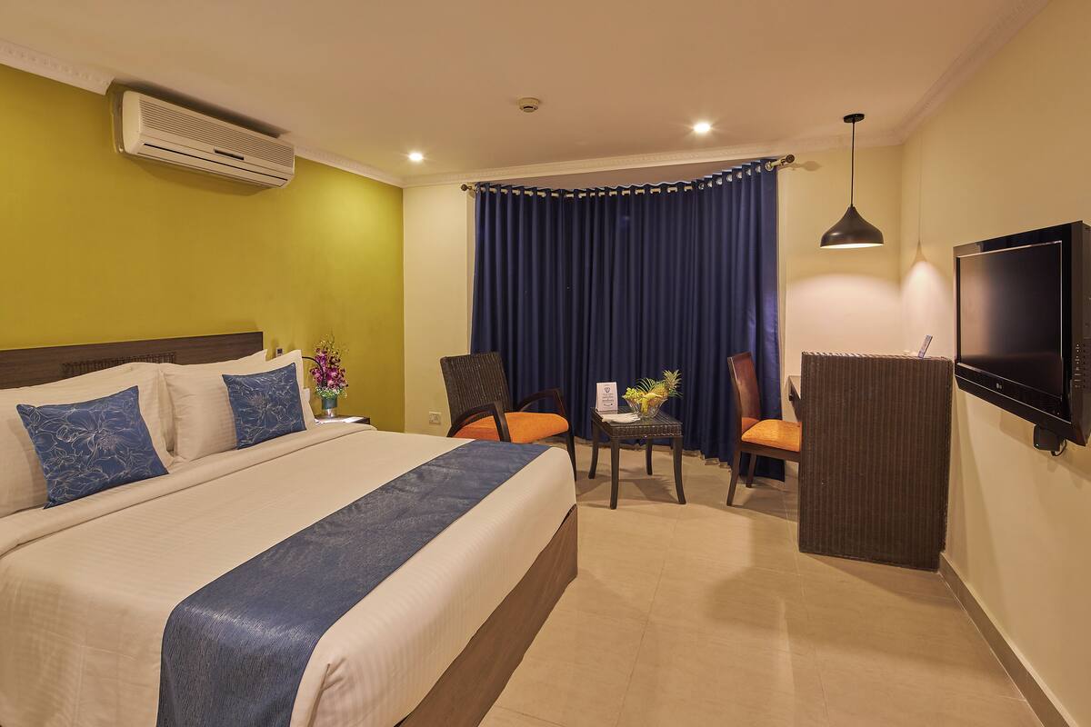A hotel room with a large bed, two chairs, a small table, a TV, and an air conditioning unit.