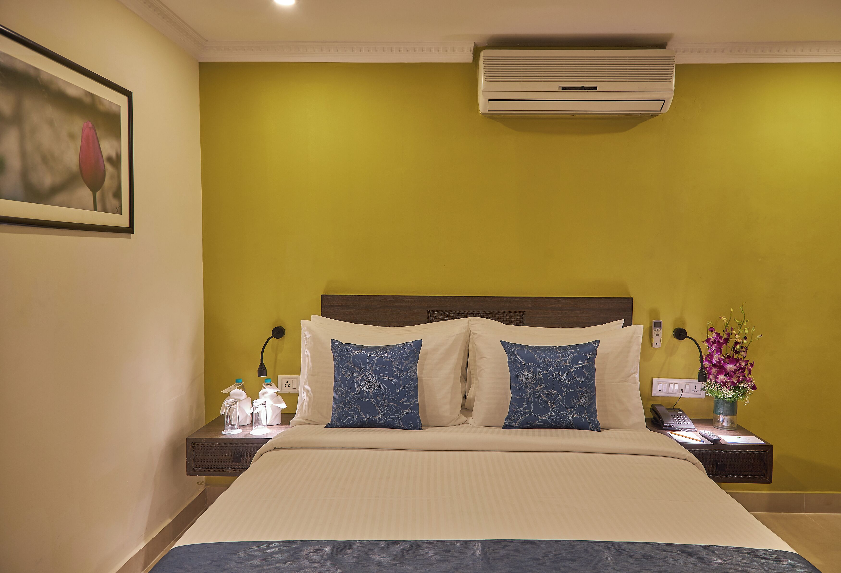 Quarto de hotel com cama, duas almofadas, mesa de cabeceira com telefone e um vaso de flores, e ar condicionado na parede.