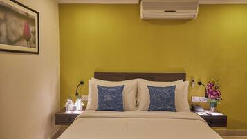 Quarto de hotel com cama, duas almofadas, mesa de cabeceira com telefone e um vaso de flores, e ar condicionado na parede.