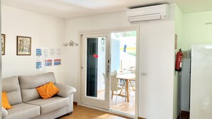 Una sala de estar con un sofá gris, un aire acondicionado blanco, una puerta de vidrio que da al exterior y una pequeña mesa con sillas.