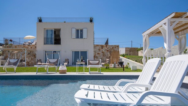 A modern pool area with lounge chairs, a stone wall, and a two-story building with balconies. - Sicily luxury Marzamemi (Noto)