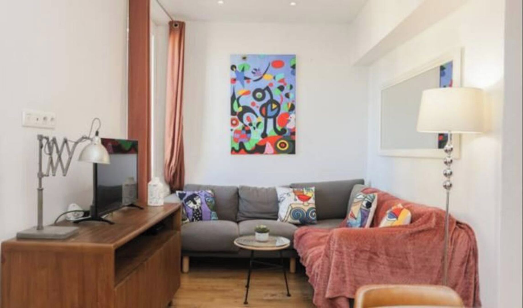A compact living space with a grey sofa, a small round table, a wooden desk, and a colorful abstract painting on the wall.