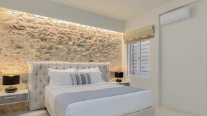A bedroom with a stone wall, a bed with white bedding, two bedside tables with lamps, and a window with blinds. - THIRAMENOUS RESIDENCE - SIX BEDROOM HOUSE with Rooftop Hot Tub (Athina)