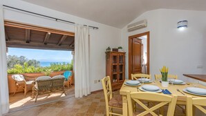 A dining area with a table set for four, a wooden cabinet, and a view through a doorway to a balcony with plants and a view of the sea. - Villetta Franca 100m From The Beach (Golfo Aranci)