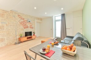 A modern living room with a brick wall, a flat-screen TV, a wooden coffee table, and a dining area with a table set for breakfast. - Premium 1  Apartment (Dijon)