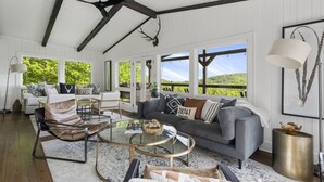 A spacious living room with a large gray sofa, a round glass coffee table, and a leather armchair. There are multiple windows offering a view of the outdoors. - High Noon: Modern Mountaintop Home w 2 Covered Porches, Firepit | Pet Friendly (Highlands)