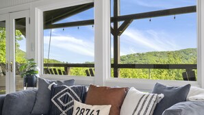 A gray sofa with multiple cushions, including a pillow with "28741 Highlands" text, positioned in front of large windows overlooking a green landscape. - High Noon: Modern Mountaintop Home w 2 Covered Porches, Firepit | Pet Friendly (Highlands)