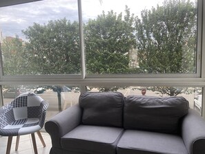 A room with a grey sofa and a patterned chair, large windows with a view of trees and buildings. - The Harbor and the Sea (Dunkerque)