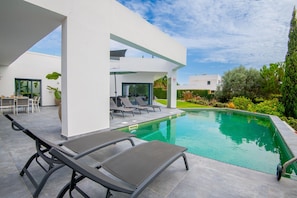 A modern outdoor pool area with lounge chairs, a dining table, and a clear view of the surrounding garden.