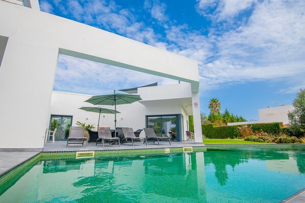A modern outdoor pool area with lounge chairs and umbrellas, surrounded by a white building with glass doors. - Casa Blanca Magnificent Villa With Modern Design, walking distance to beach (Carvoeiro)