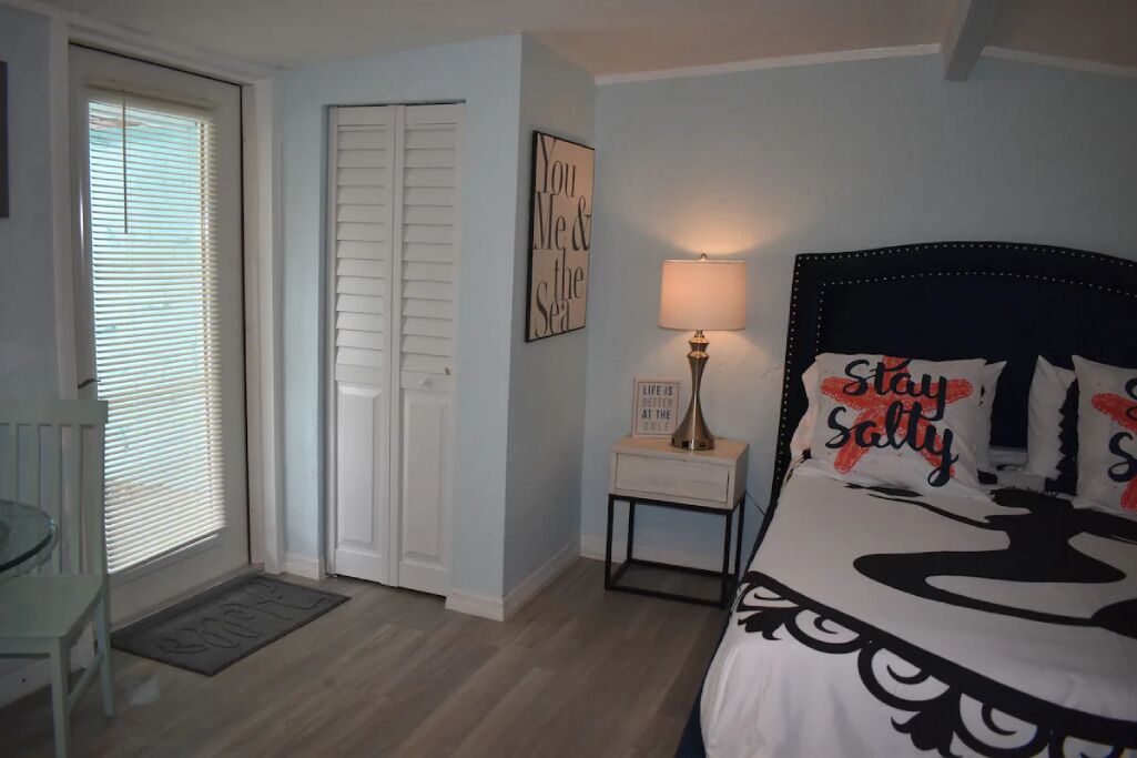 A bedroom with a bed, a bedside table with a lamp, a door, and a window with blinds.