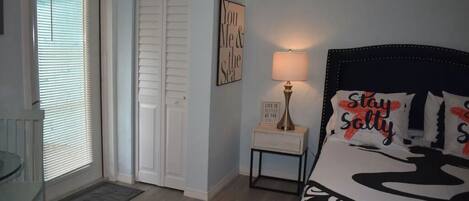 A bedroom with a bed, a bedside table with a lamp, a door, and a window with blinds.