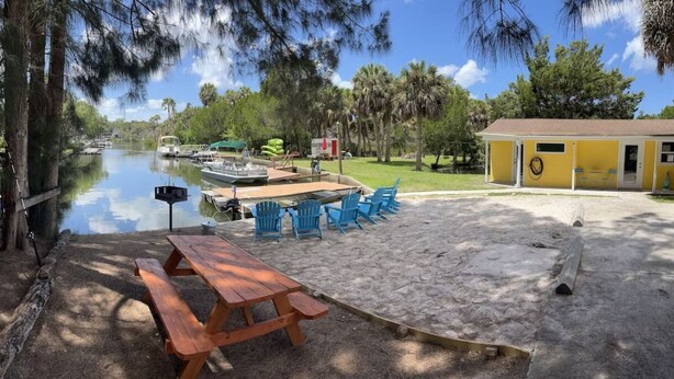 A riverside picnic area with a wooden table and blue chairs, a small yellow building, and boats docked nearby. - "Salty Siren" Mermaid Themed Room at Weeki Wachee Waterfront Adventure Retreat (Weeki Wachee)