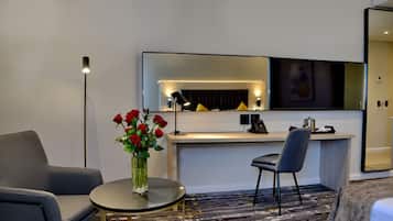 Une chambre d’hôtel moderne équipée d’un bureau, de deux chaises, d’une table ronde ornée d’un vase de roses rouges, d’une lampe sur pied et d’un grand miroir.