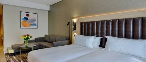 A hotel room with a large bed, a sofa, a small table with a vase of flowers, and a wall-mounted lamp.