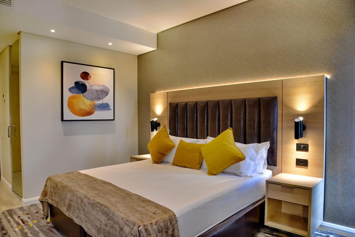 Een moderne hotelkamer met een groot bed, een hoofdbord met ingebouwde verlichting, een nachtkastje en een aan de muur bevestigd kunstwerk.