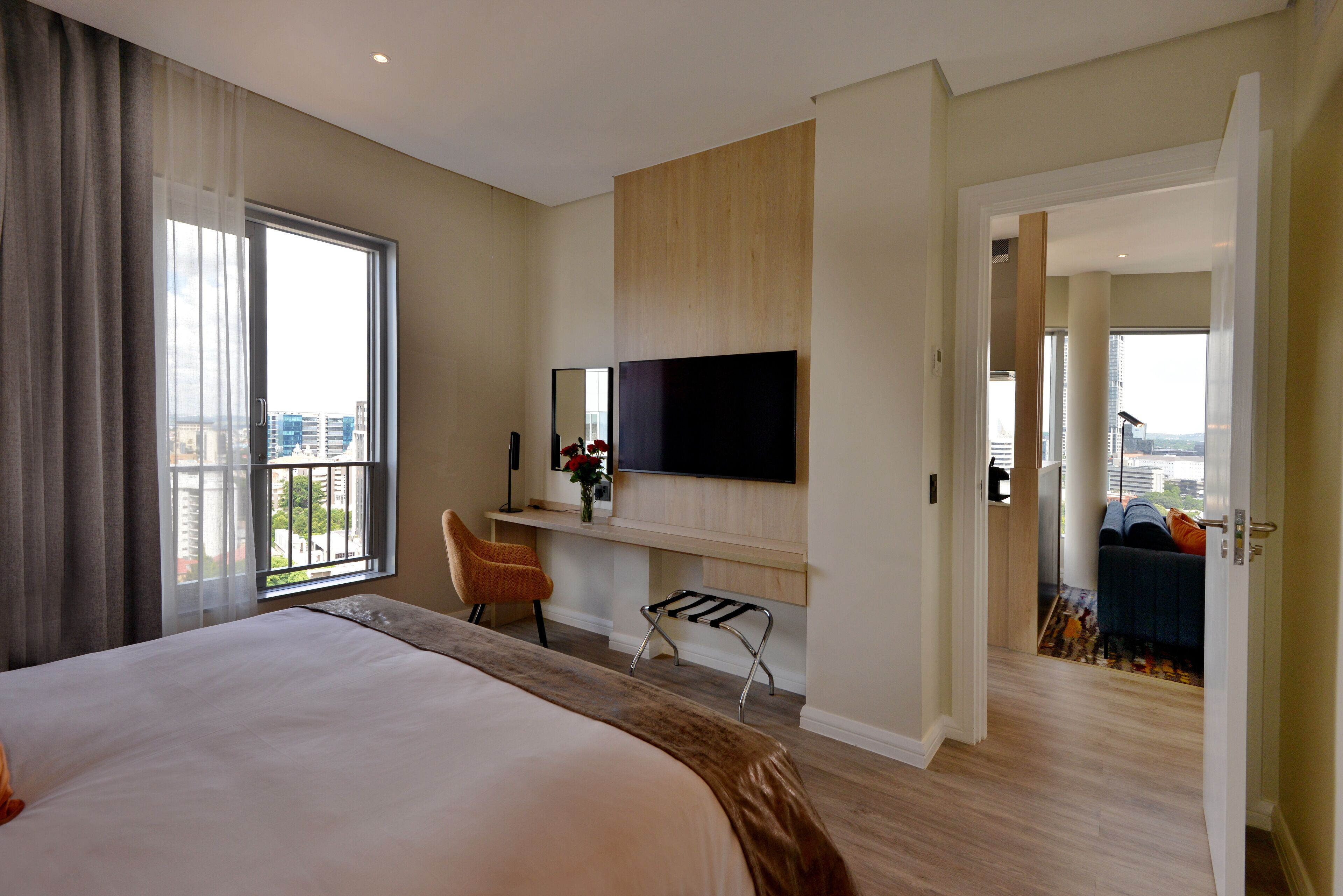 A hotel room with a large bed, a desk with a flat-screen TV, a chair, and a view of the city through a large window.