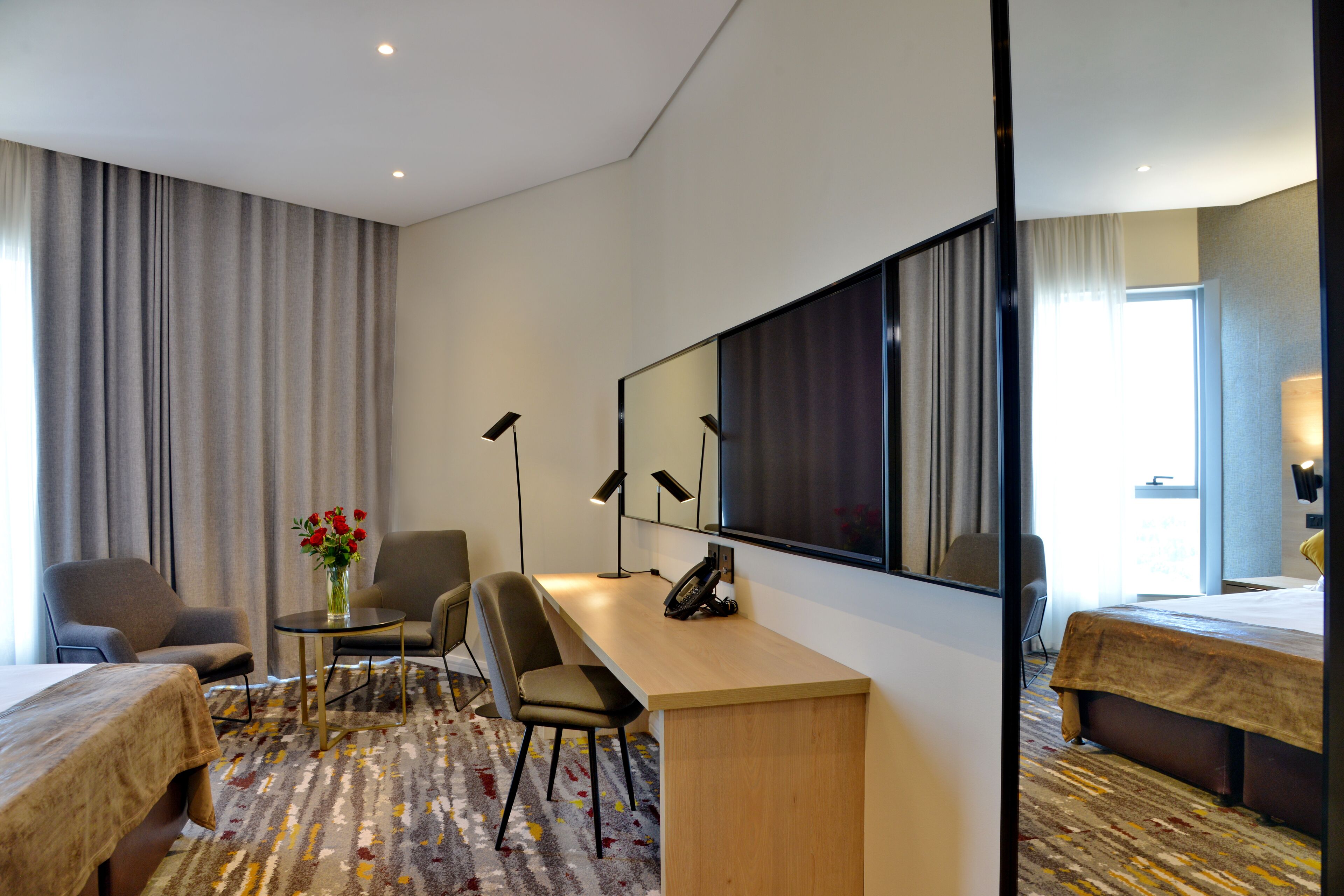 Habitación de hotel con cama, escritorio, dos sillas, una mesita con un florero, un espejo grande y piso alfombrado.