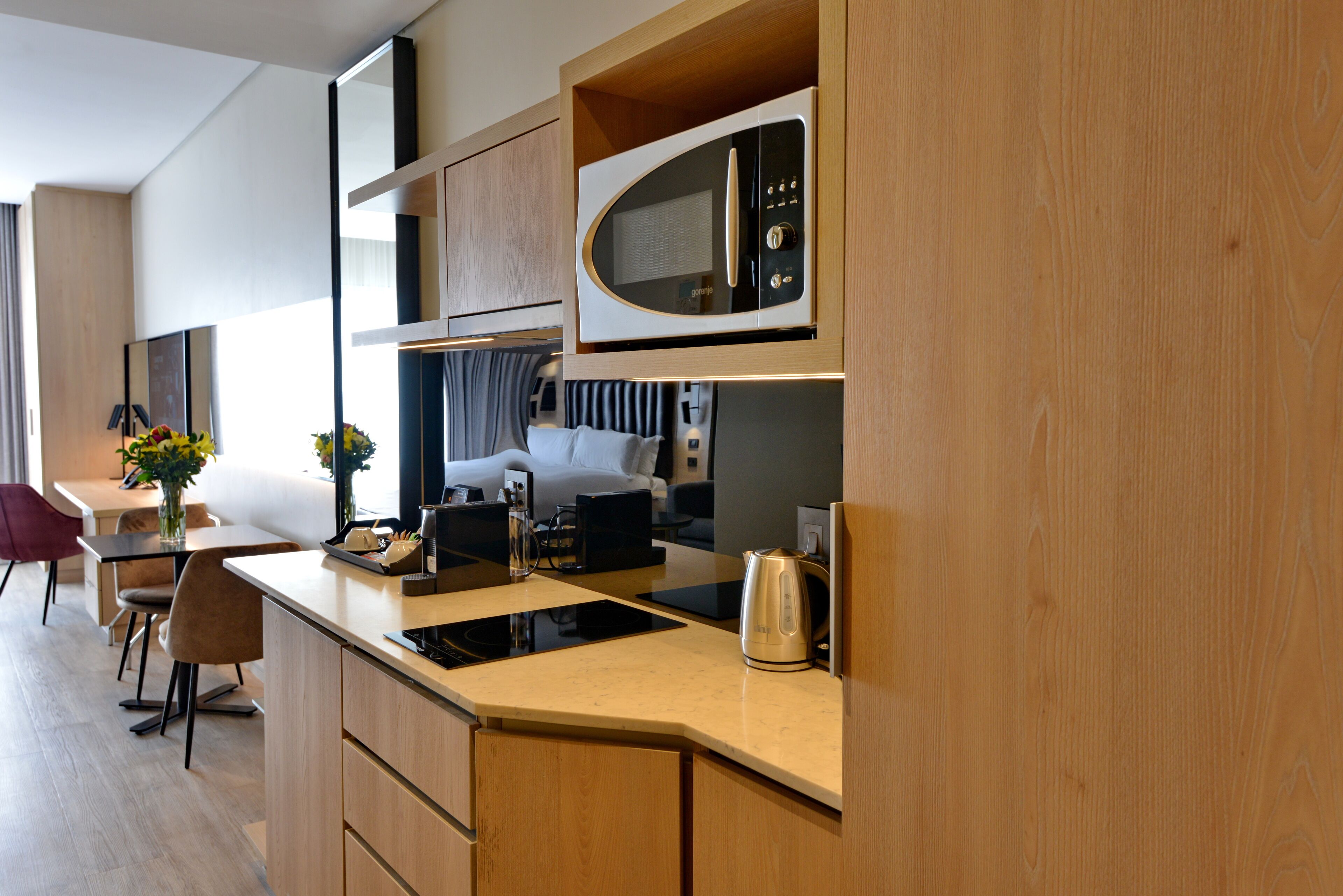 A modern kitchen with wooden cabinets, a built-in microwave, a kettle, and a dining area with a table and chairs.