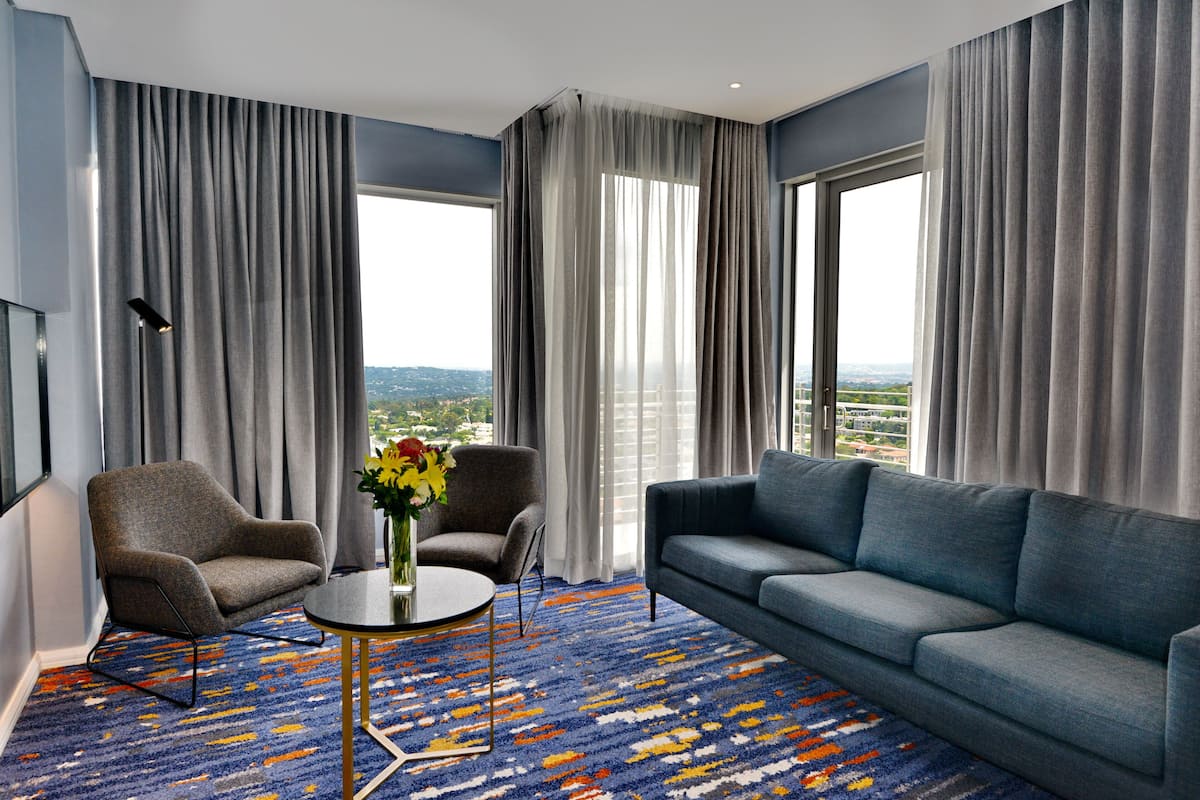 Een moderne hotelkamer met een blauwe bank, twee fauteuils, een ronde salontafel en uitzicht op een stadsbeeld door grote ramen.