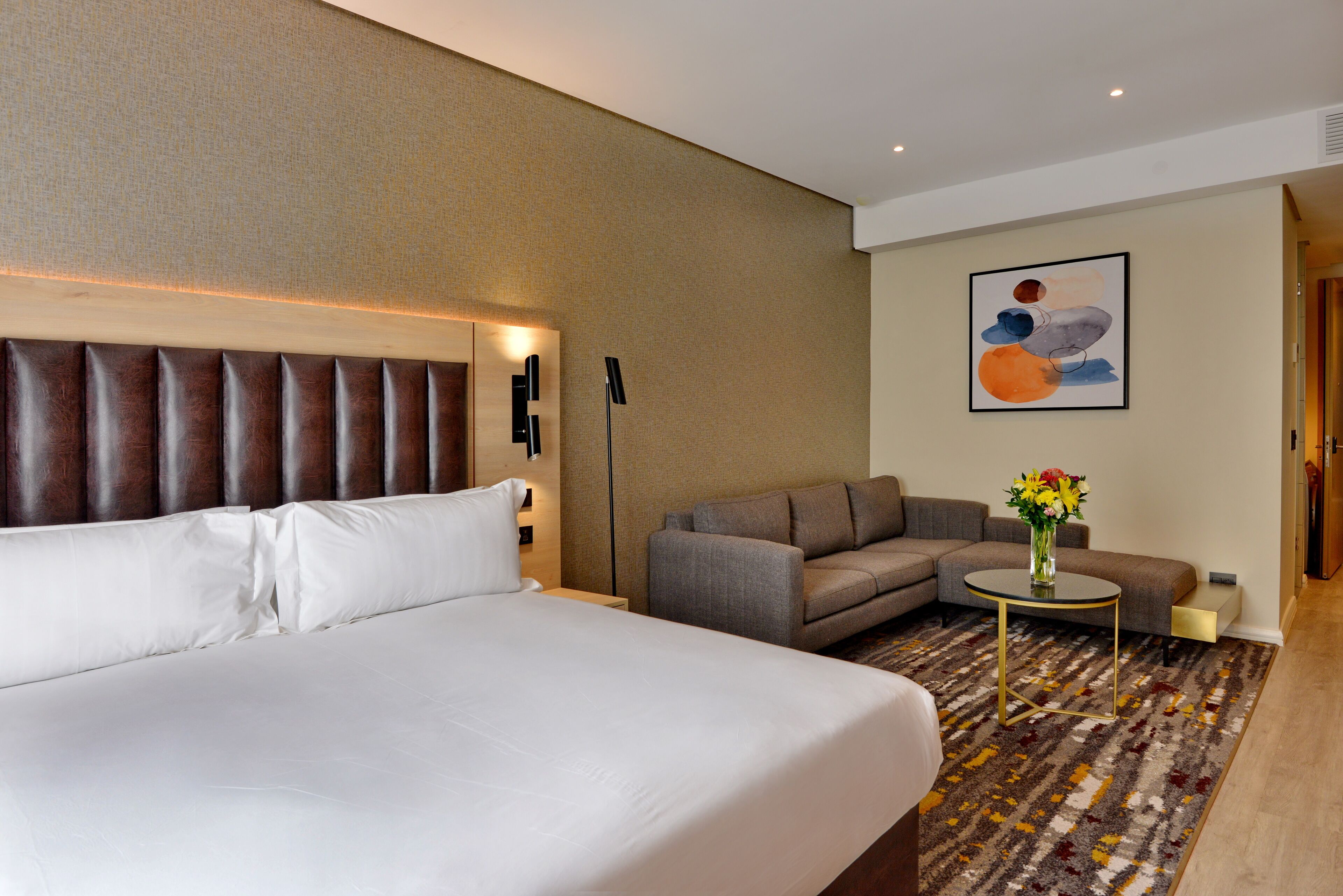 Een hotelkamer met een groot bed, een bank, een kleine ronde tafel met een bloemenvaas, en een schilderij aan de muur.