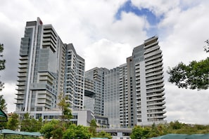 Um edifício moderno alto com várias varandas, rodeado por vegetação e sob um céu nublado.