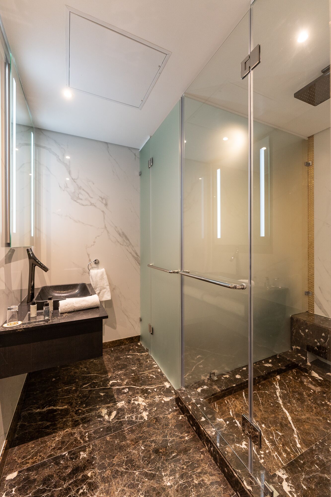 Un baño moderno con cabina de ducha de vidrio, suelo de baldosas de mármol y lavamanos negro con espejo.
