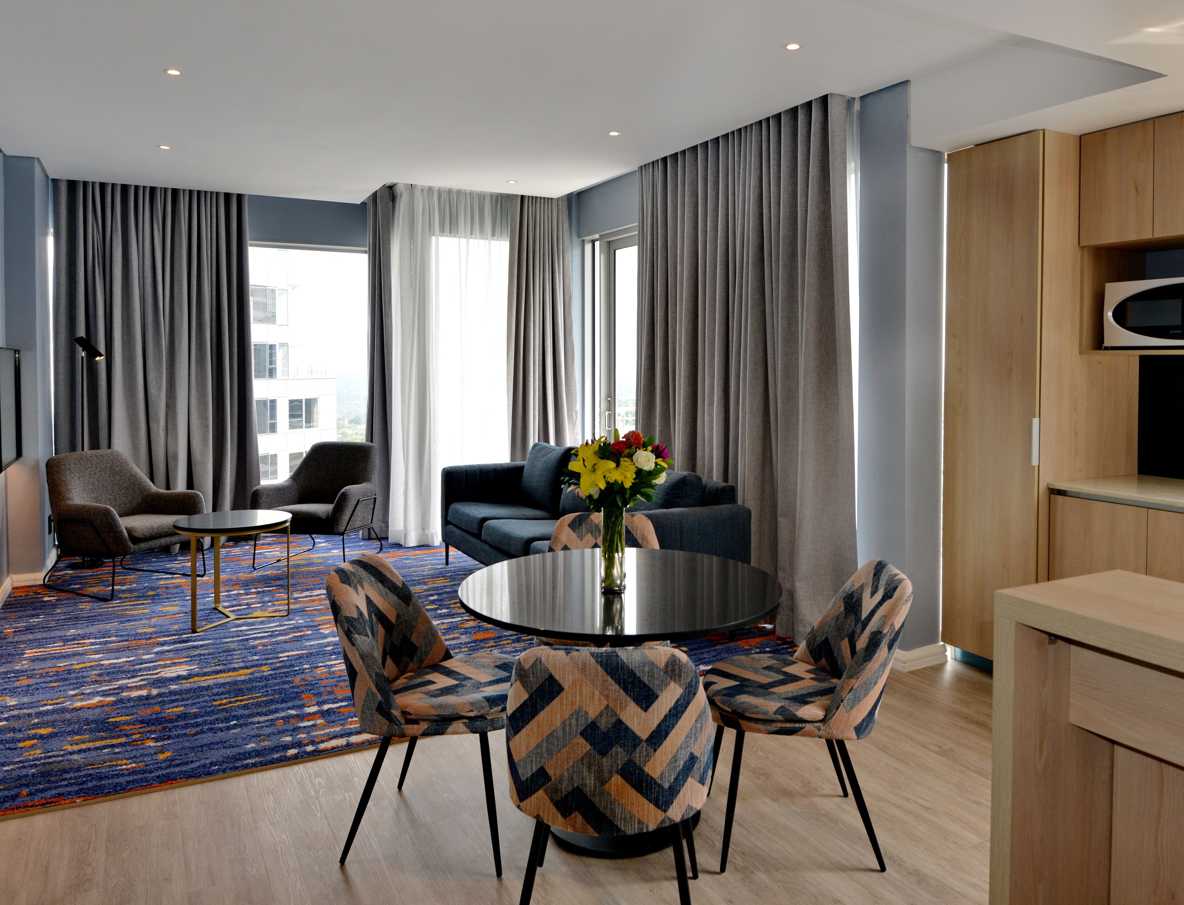 Ein modernes Hotelzimmer mit großem Fenster, einer Sitzecke, zwei Sesseln, einem Couchtisch und einem Essbereich mit gemustertem Teppich.