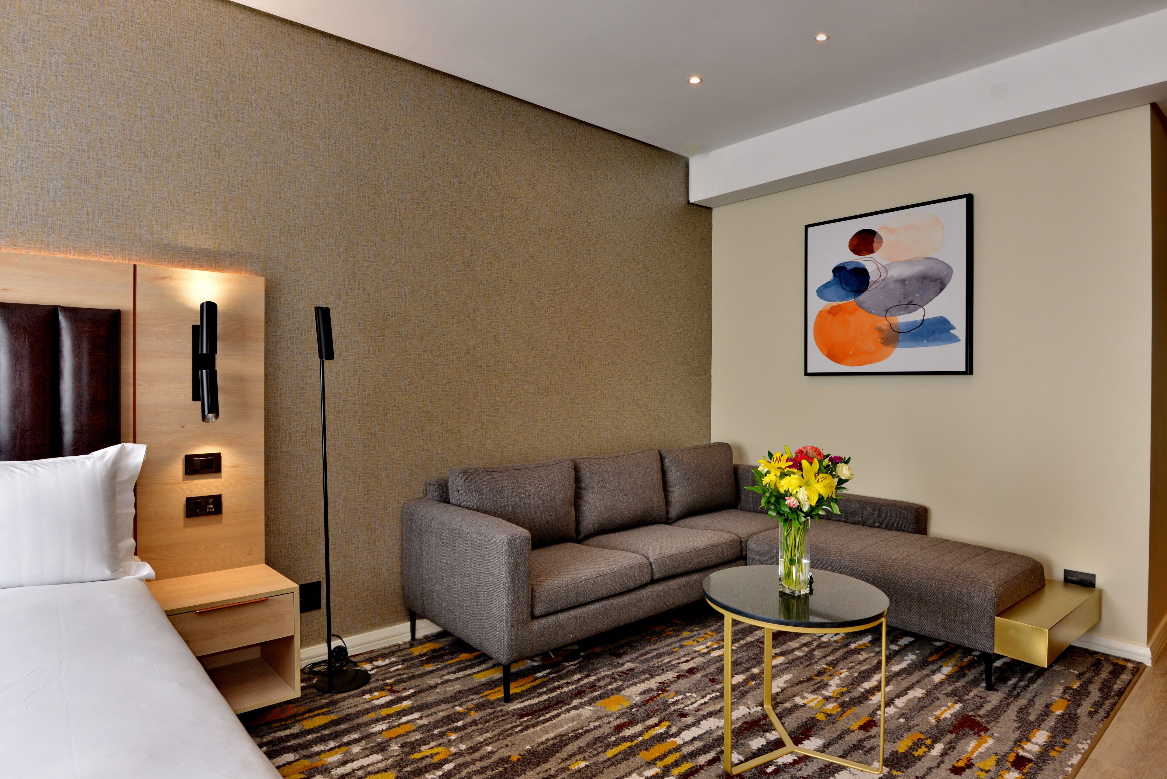 A modern hotel room with a sofa, a small table with a vase of flowers, a bedside table, and a floor lamp.