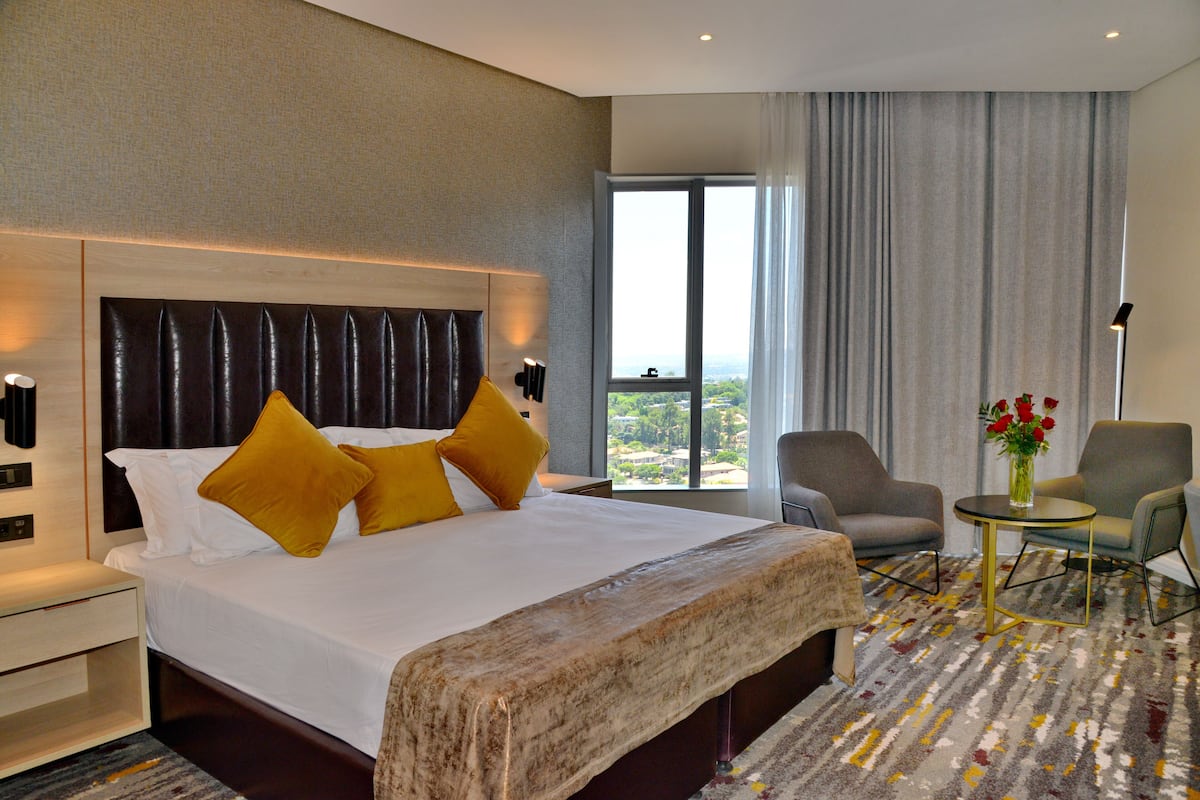 Een hotelkamer met een groot bed, twee fauteuils, een tafeltje met een bloevas en uitzicht op de stad door het raam.