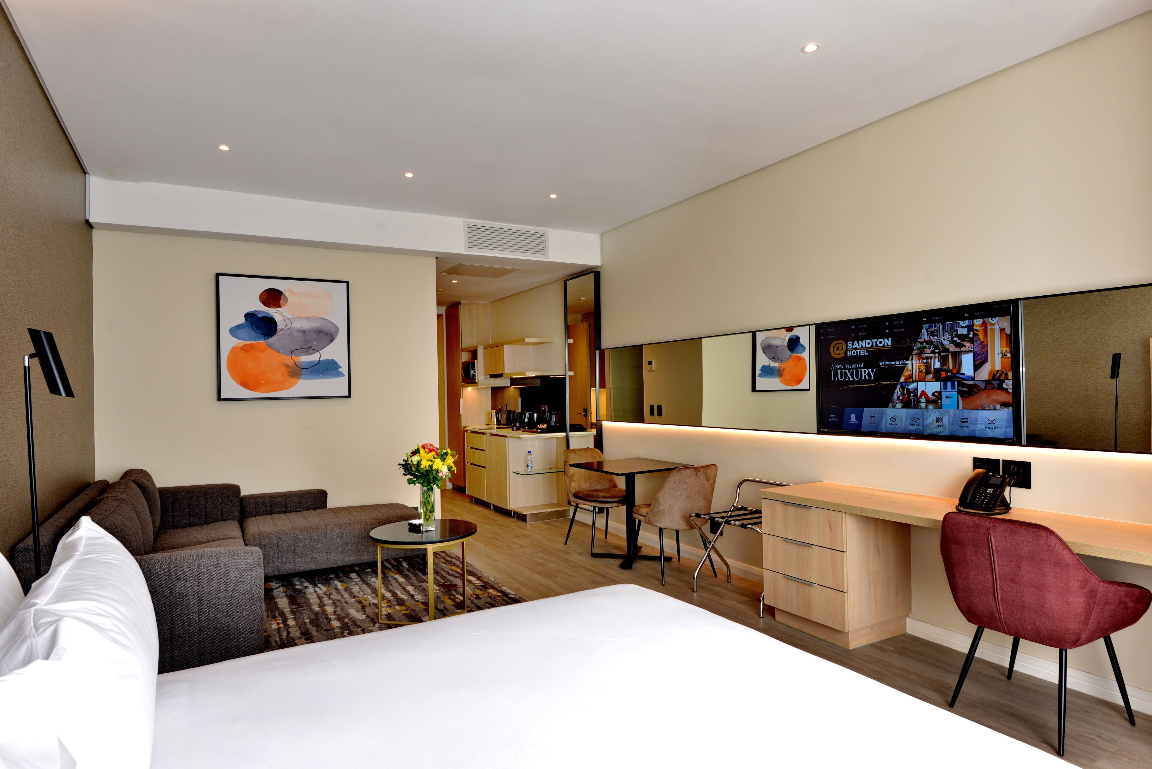 Une chambre d’hôtel moderne dotée d’un grand lit, d’un canapé, d’un bureau avec une chaise, d’une petite table ornée d’un vase de fleurs, d’un coin cuisine et d’un grand miroir.