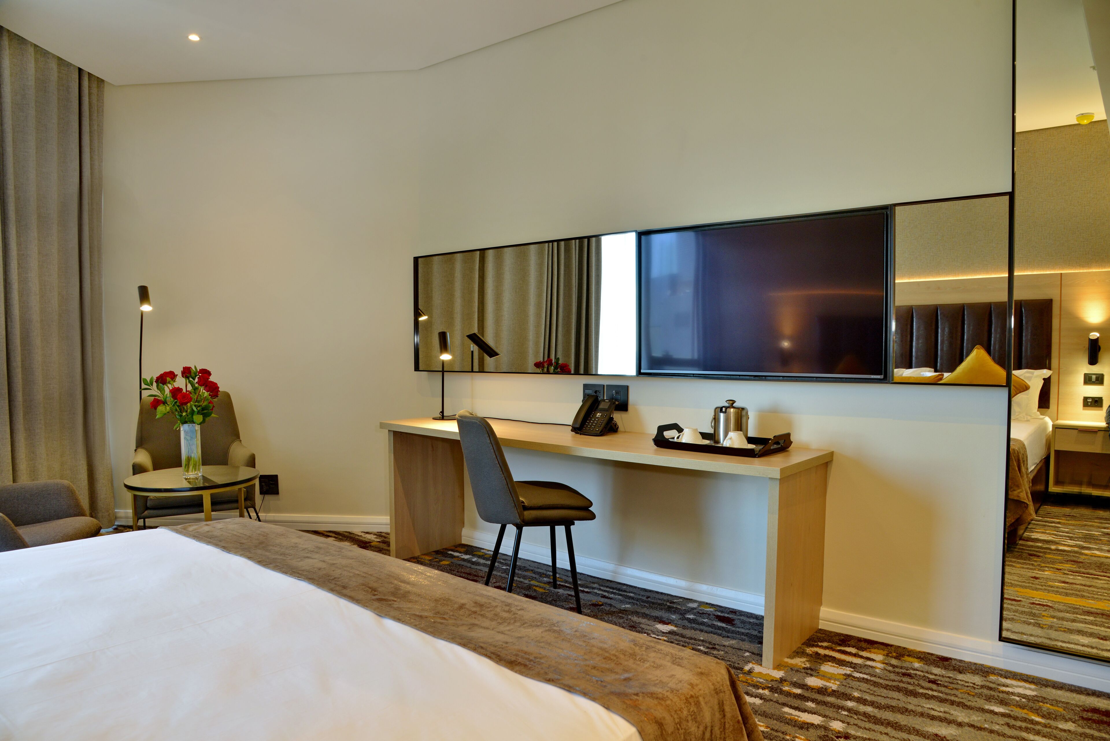 A hotel room with a large bed, a desk with a chair, a mirror, and a small table with a vase of flowers.