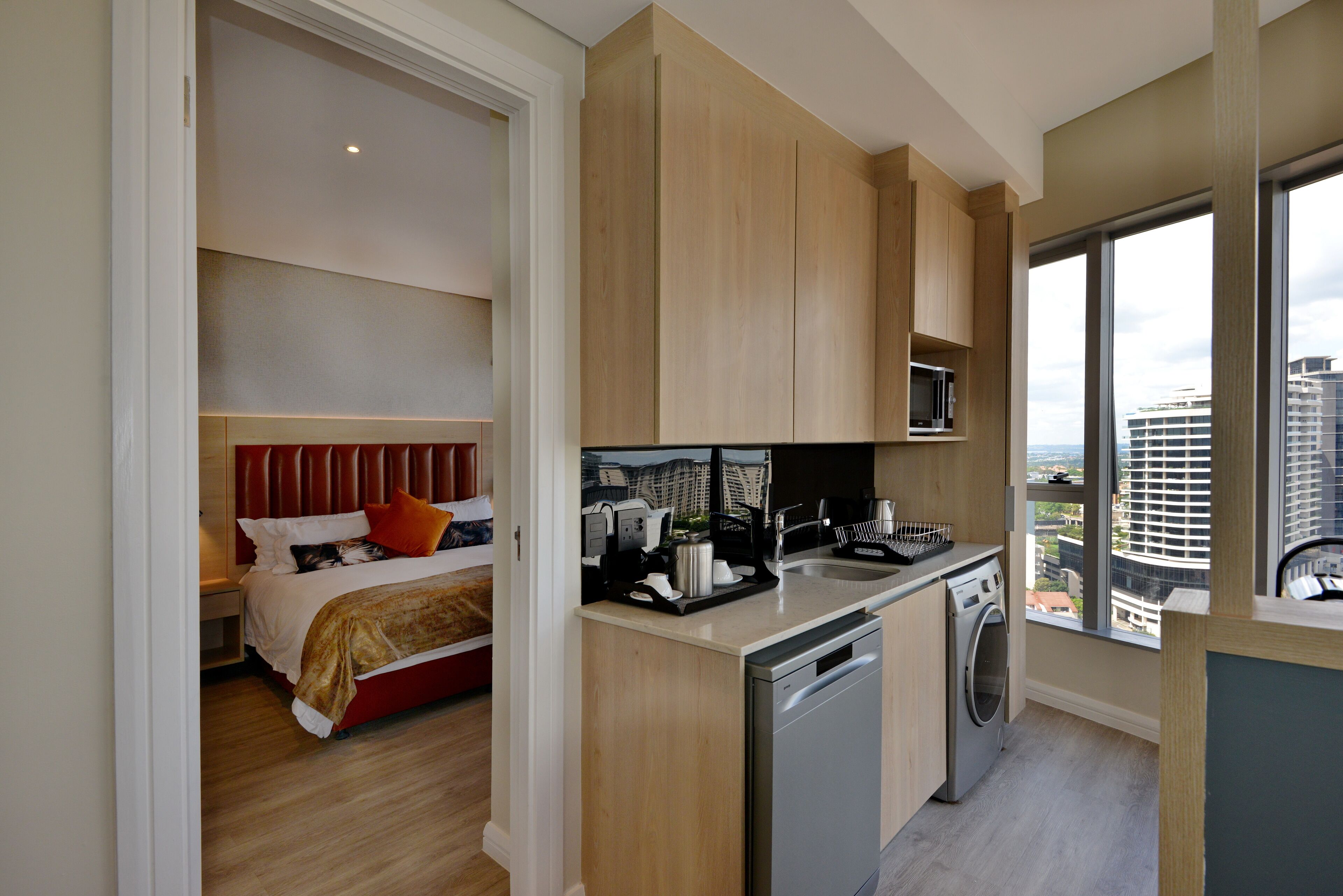 Habitación de hotel moderna con cama, zona de cocina con lavavajillas y un amplio ventanal con vistas a la ciudad.
