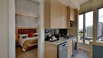Habitación de hotel moderna con cama, zona de cocina con lavavajillas y un amplio ventanal con vistas a la ciudad.